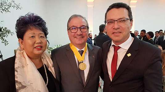 Três pessoas (Dra. Margarete Kishi de blusa preta e lenço, Dr. Francisco Caravante (de terno e medalha com faixa amarela e Dr. Marcos Machado, de terno e gravata vermelha) estão juntos na premiação 