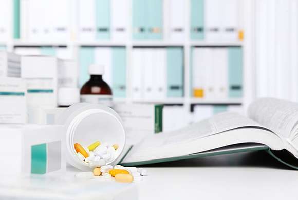 Imagem ilustra uma bancada em uma farmácia com medicamentos ao fundo. Sobre a bancada, um frasco de medicamentos está deitado com alguns comprimidos espalhados ao lado de um livro aberto.