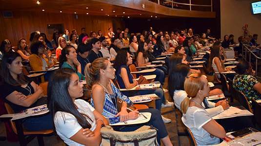Farmacêuticos e estudantes de Farmácia (homens e mulheres de diferentes etnias, majoritariamente jovem) sentados em um grande auditório de universidade assistem a palestras