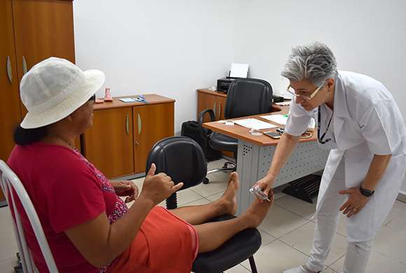 Pessoa sentada em cadeira com a perna esticada e farmacêutica verificando sensibilidade dos pés da paciente 