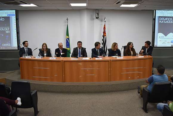 Foto de uma mesa localizada na frente de um grande auditório composta por homens e mulheres: nas laterais há dois telões e ao fundo as bandeiras do Brasil e do Estado de São Paulo