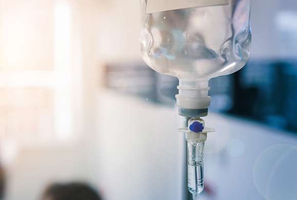 Imagem em primeiro plano em que se vê um frasco de soro suspenso tendo ao fundo em ambiente tipicamente hospitalar