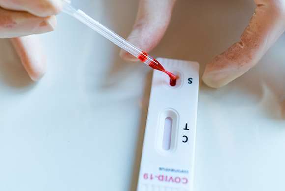 Foto de um dispositivo de teste rápido em primeiro plano nas mãos de um profissional da saúde cujas mãos, com luvas de látex branca, insere um pingo de sangue para fins de testagem