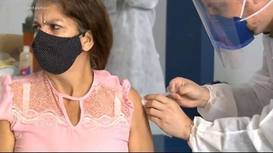 profissional paramentada com avental branco e máscara aplica vacina em braço de mulher de cabelos castanhos 