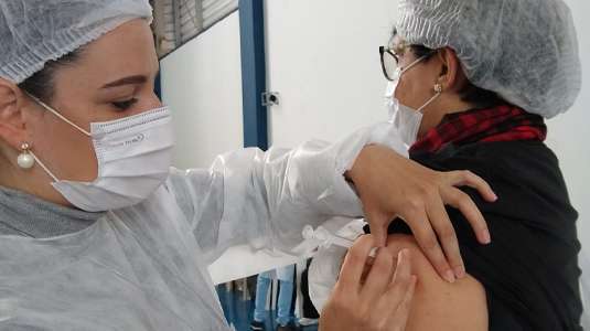 Farmacêutica com touca, máscara e avental brancos aplica vacina no braço de mulher de óculos e camiseta preta