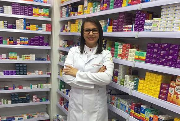 Farmacêutica de jaleco branco, braços cruzados, ela usa óculos, tem cabelo castanho na altura dos ombros e está em frente a uma parede com caixas de medicamentos 
