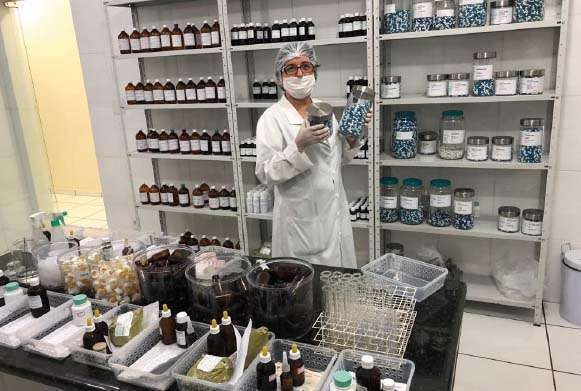 Farmacêutica com jaleco branco, máscara e touca em pé segurando um pote de vidro com medicamentos. Atrás uma prateleira com medicamentos (cápsulas) em potes de vidro e à frente uma bancada com matérias-primas para a produção de fitoterápicos