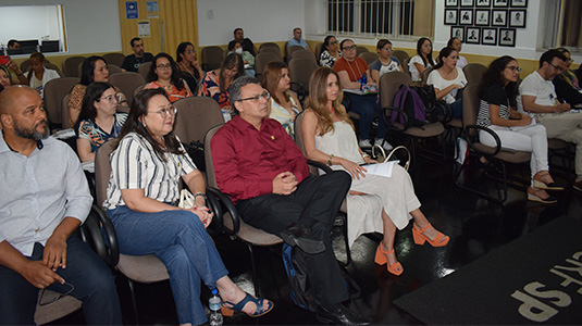 Foto de um auditório repleto de pessoas sentadas acompanhando um evento. O público é formado por homens e mulheres de diversas idades, etnias e composições corporais diferentes