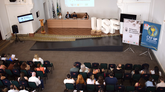 Vista de cima de auditorio lotado e uma mesa à frente em cima do palco com quatro pessoas