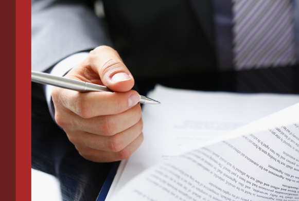 Imagem mostra a mão de uma pessoa segurando uma caneta e um documento sobre a mesa