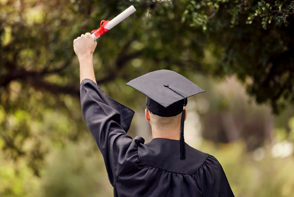 Foto mostra acadêmico de costas com roupa de formatura e levantando o seu diploma