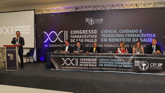 Uma pessoa em pé e seis pessoas sentadas durante a cerimônia de abertura do XXII Congresso Farmacêutico de São Paulo 
