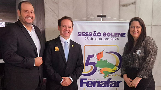 Foto de três pessoas posadas, sendo dois homens e uma mulher. Os dois homens vestem terno preto e camisa branca, um deles é alto e possui compleição mais forte. Ambos são brancos, calvos e os cabelos ralos são castanhos escuros. A mulher é branca, tem cabelos pretos longos e lisos. Veste camisa estampada e saia na cor escura. Por tràs deles há um banner onde se lê "sessão solene 50 anos - Fenafar"